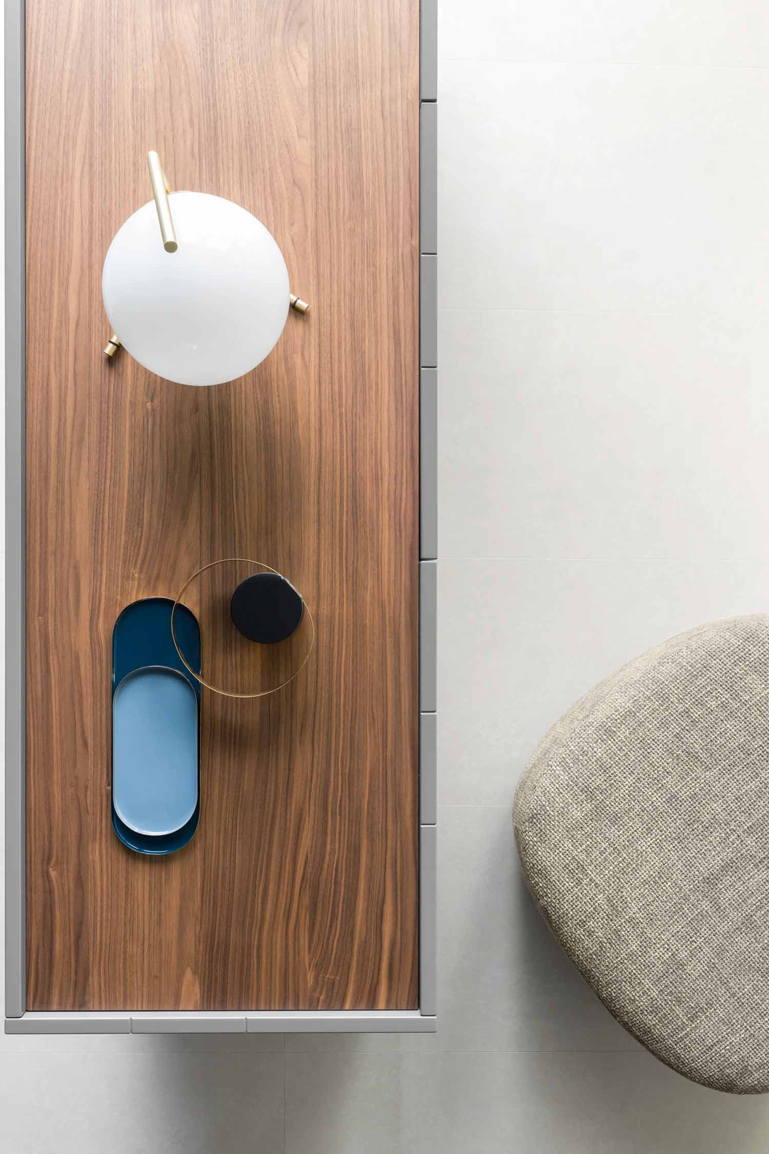 An overhead shot captures a minimalist wooden console table with various decorative objects arranged on its surface. A round white globe lamp with brass accents is at the top. Below it, a bright blue oval-shaped tray holds a thin gold oval, and to its right, a small black circular object with a gold ring is placed. The table has a light grey frame. To the right, a textured beige upholstered stool or seat is partially visible against a light grey floor.