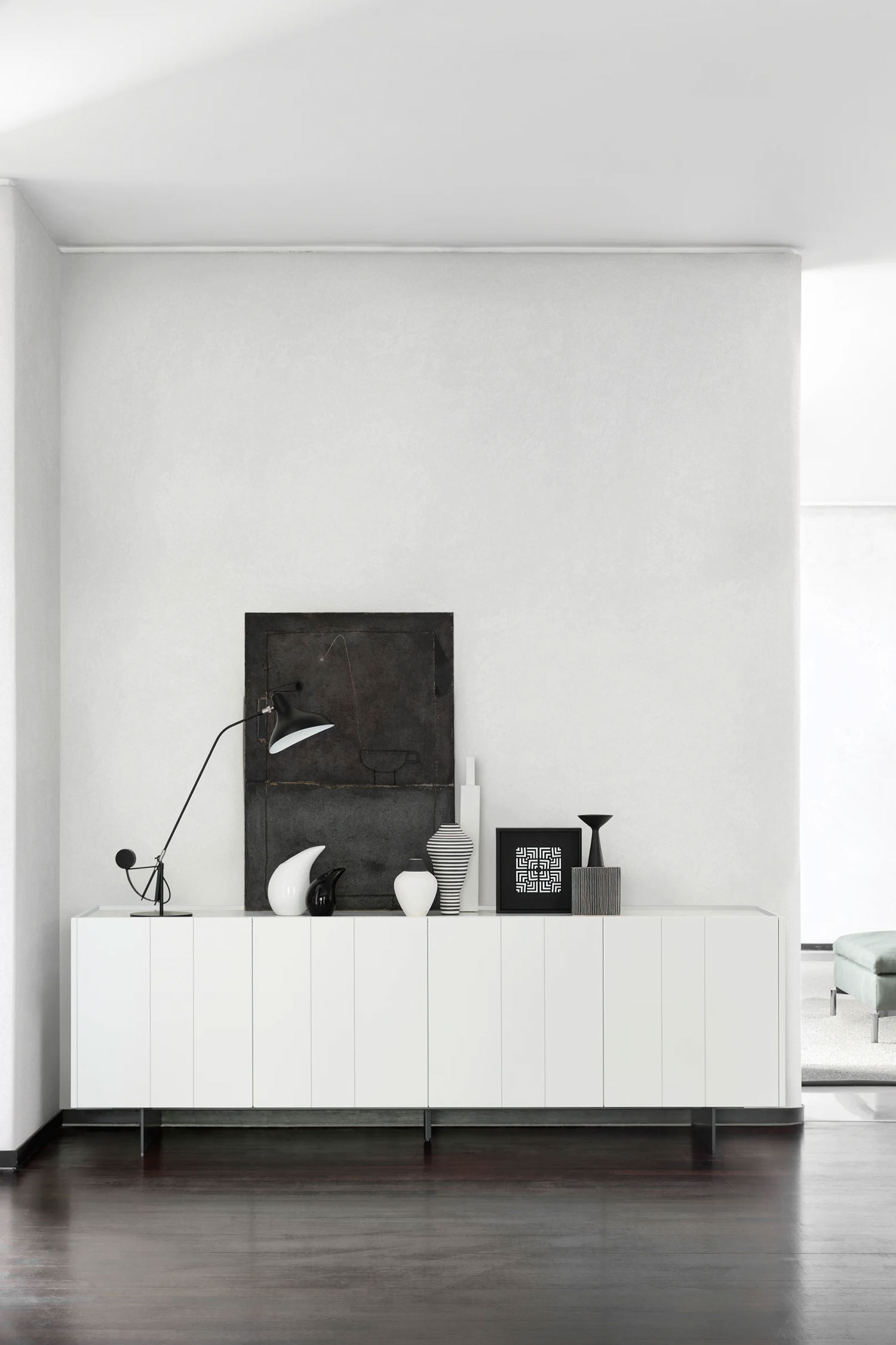 A full-view shot of a modern, minimalist white sideboard with six vertically aligned doors and thin metal legs. It is positioned against a clean white wall. The top surface of the sideboard is styled with a variety of decorative objects, including a tall black desk lamp, a black abstract painting leaning against the wall, and several vases in black, white, and striped patterns. A small black and white geometric artwork and a black vase with a triangular base are also present on the far right. The floor is made of polished dark wood.