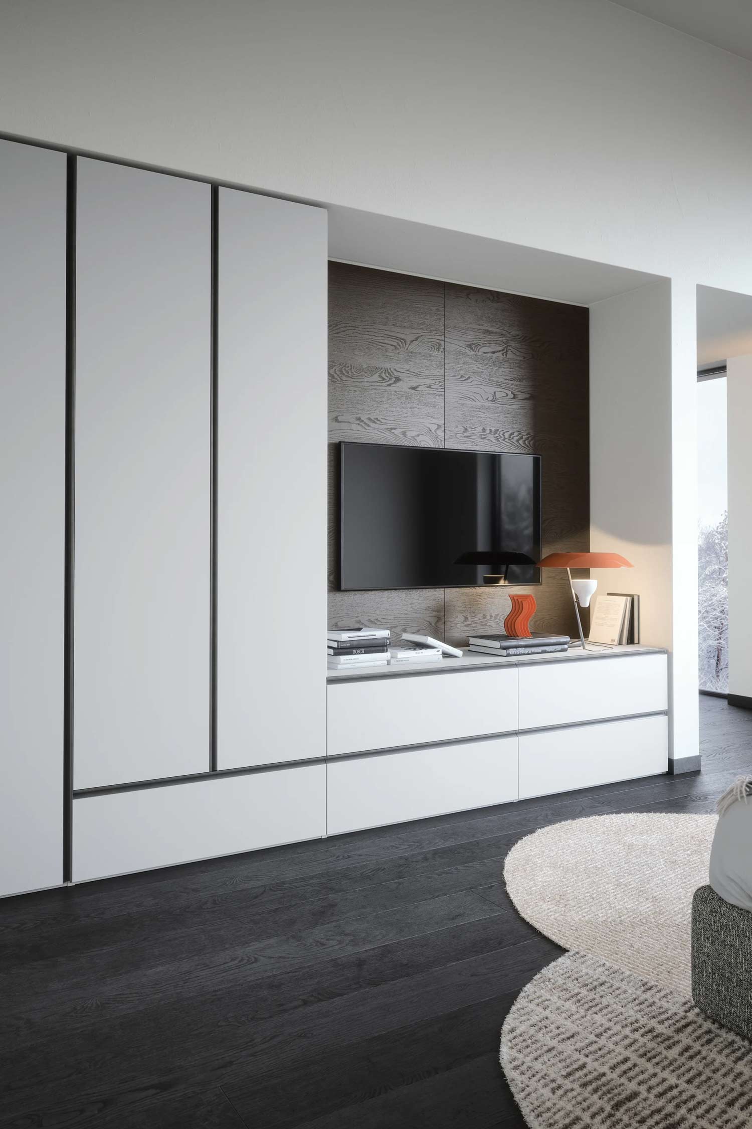 A close-up of a modern media unit with a television mounted on a dark wood paneled wall. The unit below has a white base with multiple drawers and a long shelf holding books and a unique orange lamp. The unit is integrated with tall, white wardrobe panels on the left.