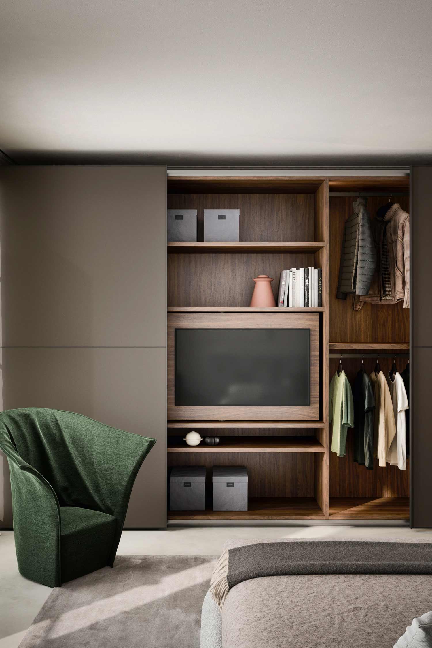 A close-up of a modern built-in closet and media unit in a bedroom. The unit features sliding dark brown doors, with one section open to reveal a television centered on a wood shelf. A separate section on the right shows hanging clothes. A green armchair sits on a gray rug on the left.