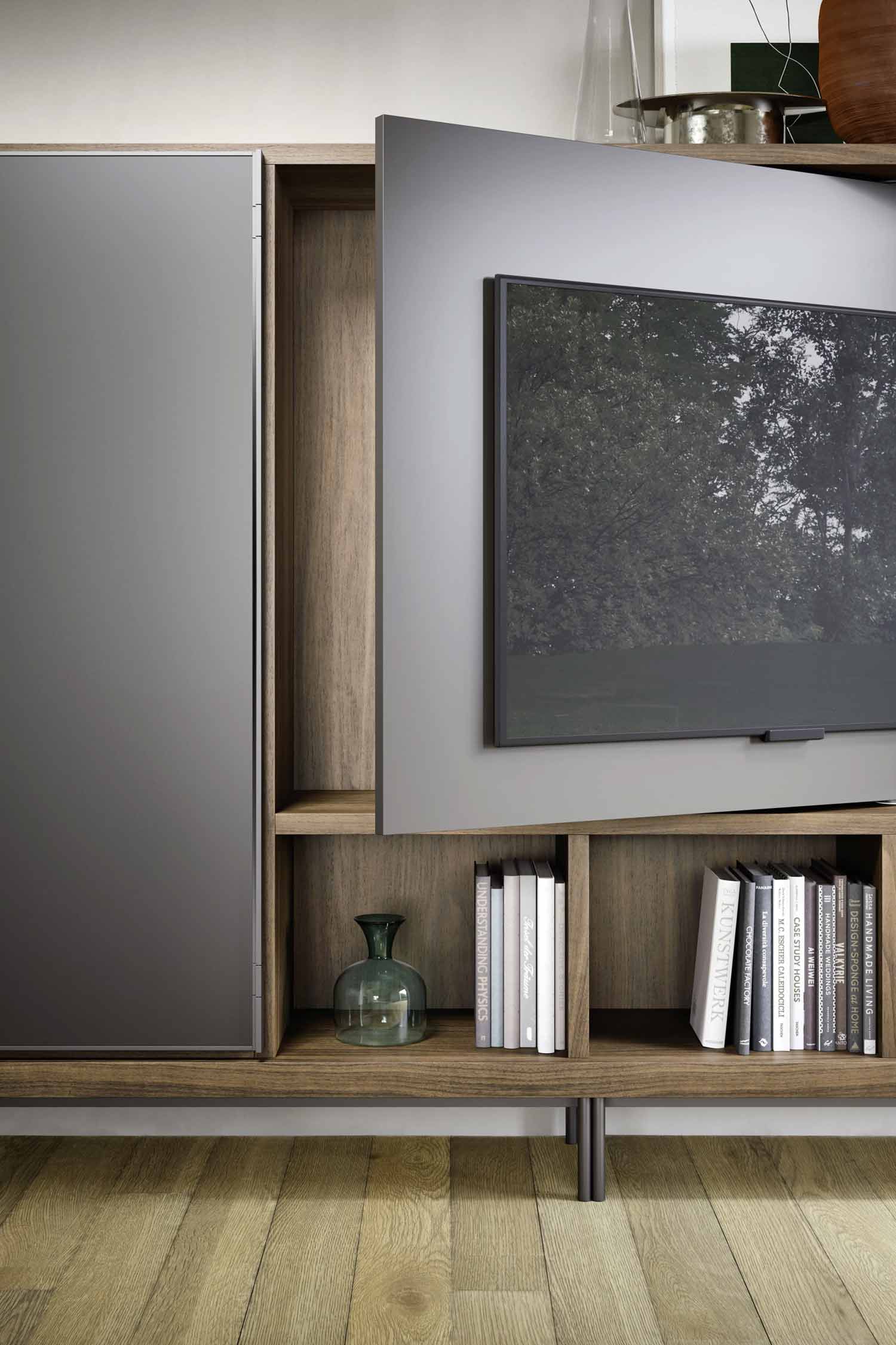 A close-up shot of a modern media unit, showing a television integrated into a gray and wood bookshelf. The gray panel for the TV can slide to the side, revealing a hidden compartment. The shelves below hold a green vase and a stack of books.