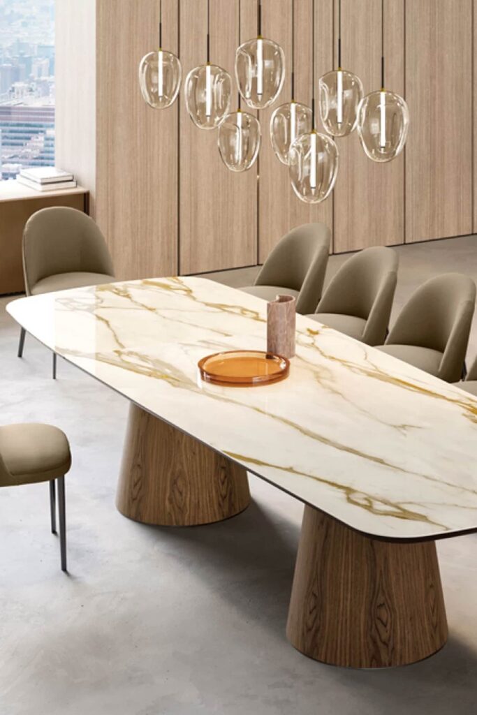 A close-up, angled view of the Teo dining table's luxurious top, featuring a white surface with prominent gold veining that mimics natural marble. The table is supported by two wide, truncated wood cone bases. The scene is lit by a modern cluster of glass pendant lights and includes tan upholstered dining chairs.