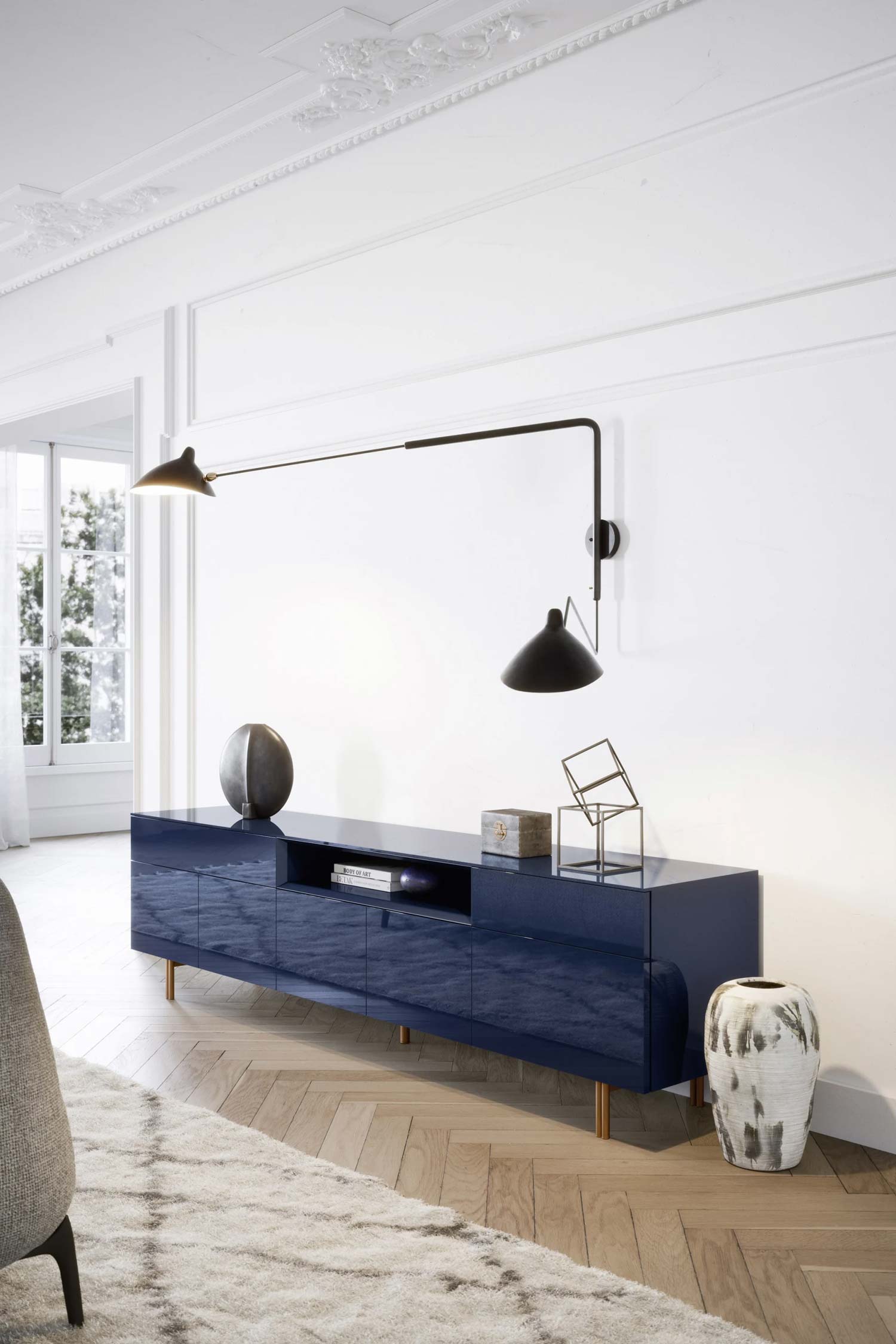 A stylish living room featuring a long, glossy dark blue media console on thin, copper-colored legs. On top of the cabinet are two decorative objects—a large, dark sphere and a small, gold-toned geometric sculpture. A long, black articulated wall lamp is mounted above, casting light on the unit. To the right of the console is a tall, textured white and brown vase. The background shows a white wall with intricate molding and a large window. A light-colored, shaggy rug is in the foreground.