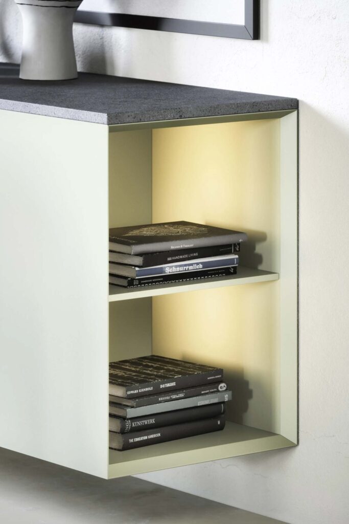 A close-up of the open shelving on a contemporary floating cabinet. The cabinet's frame is a light off-white color, and the interior of the shelves has a soft yellow glow from built-in lighting. The shelves are filled with stacks of books with various dark-colored spines. The top of the cabinet is a dark gray, textured material.