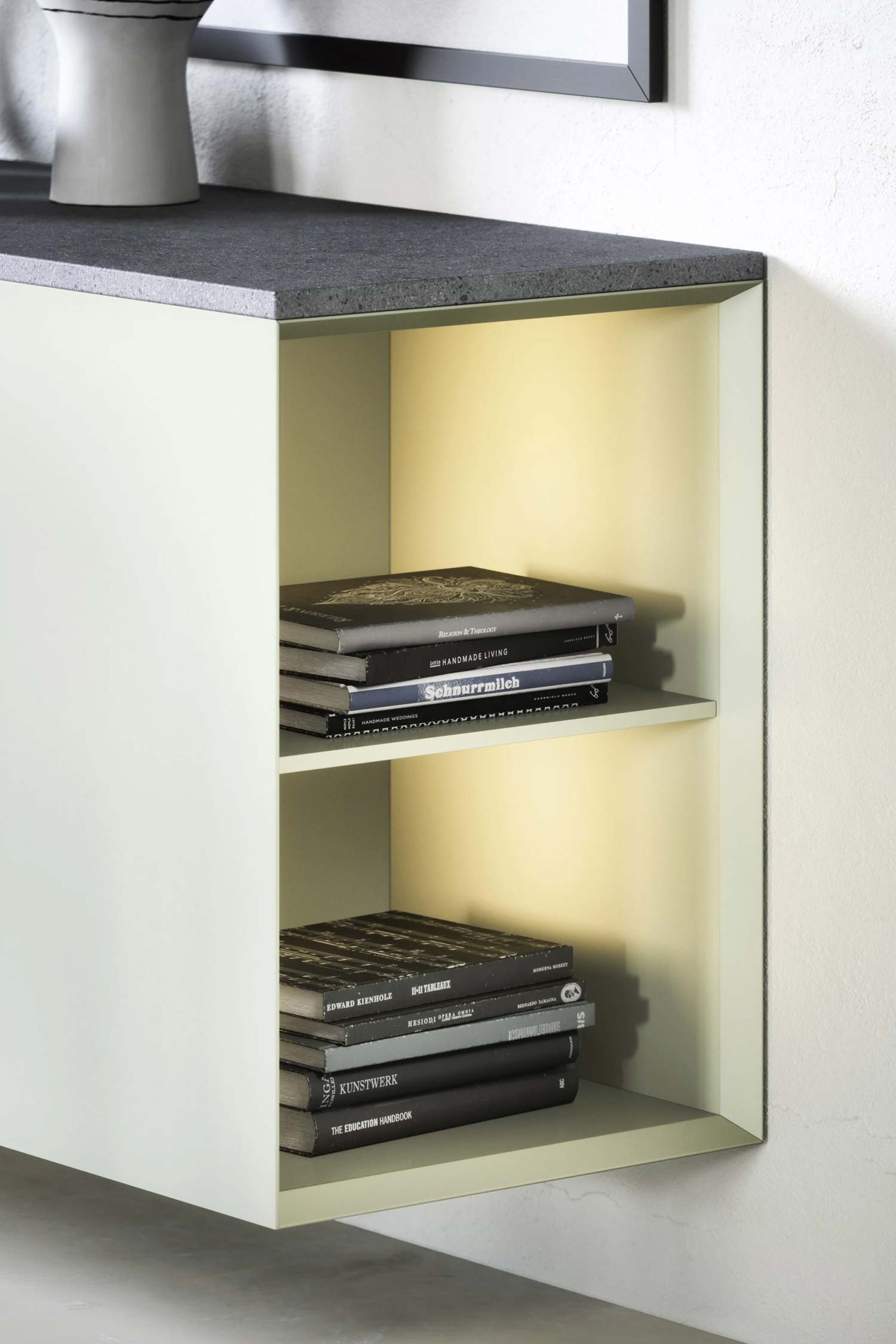 A close-up of the open shelving on a contemporary floating cabinet. The cabinet's frame is a light off-white color, and the interior of the shelves has a soft yellow glow from built-in lighting. The shelves are filled with stacks of books with various dark-colored spines. The top of the cabinet is a dark gray, textured material.