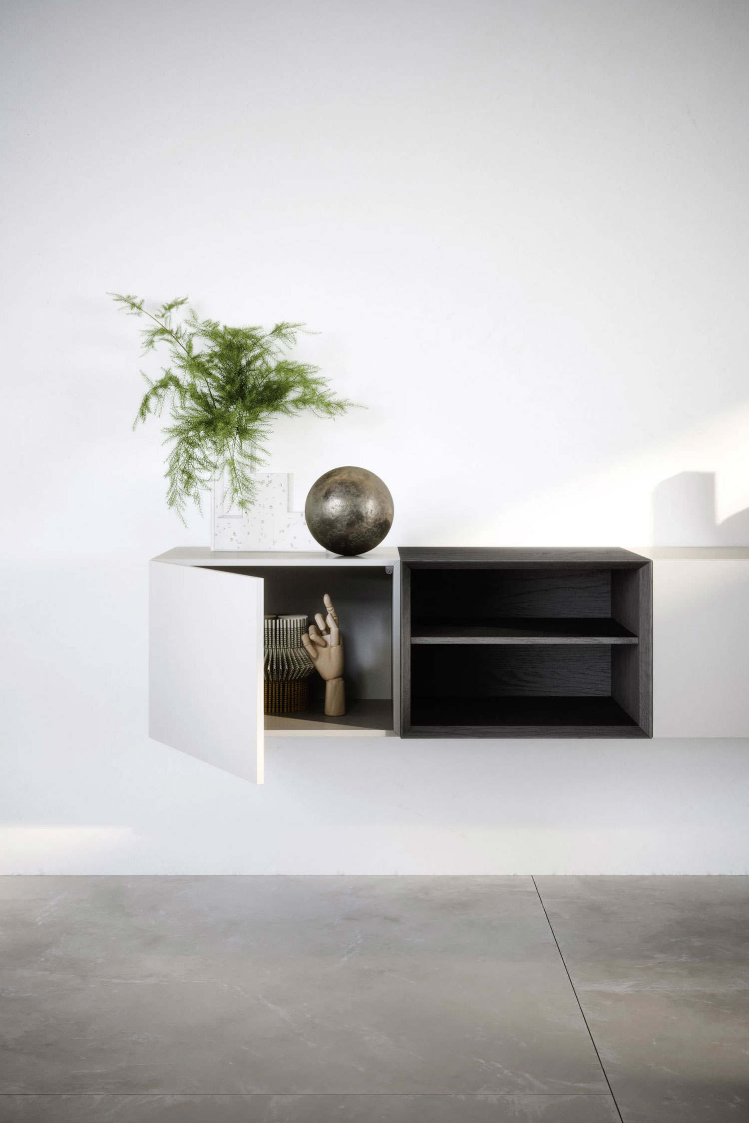 A modern, two-toned floating cabinet mounted on a white wall. The unit consists of a light off-white section with a door that is ajar, and a dark, charcoal-colored open shelving section with two shelves. Decorative objects, including a potted fern, a dark metallic sphere, and a wooden hand, are placed on top of and inside the cabinet. The floor is a light gray concrete.