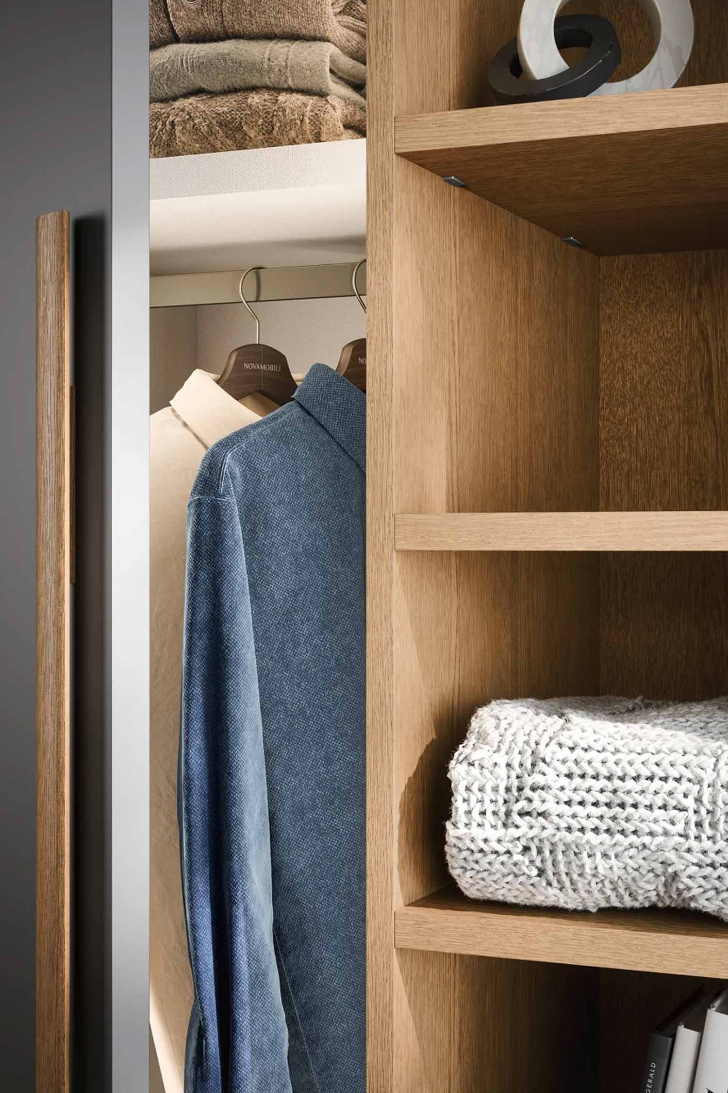 A close-up, vertical shot of the wardrobe interior. The left side shows a wardrobe door partially ajar, revealing a hanging light blue shirt. The right side shows a detailed view of the open shelving unit, which holds a folded, light-colored sweater and decorative objects on the upper shelves.