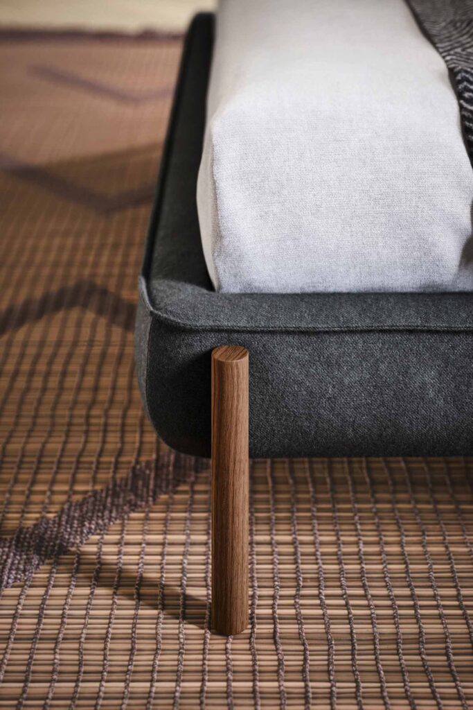 A detailed close-up of the dark gray upholstered bed frame, showing a single, cylindrical wooden leg. The bed rests on a textured, brown woven mat or rug.
