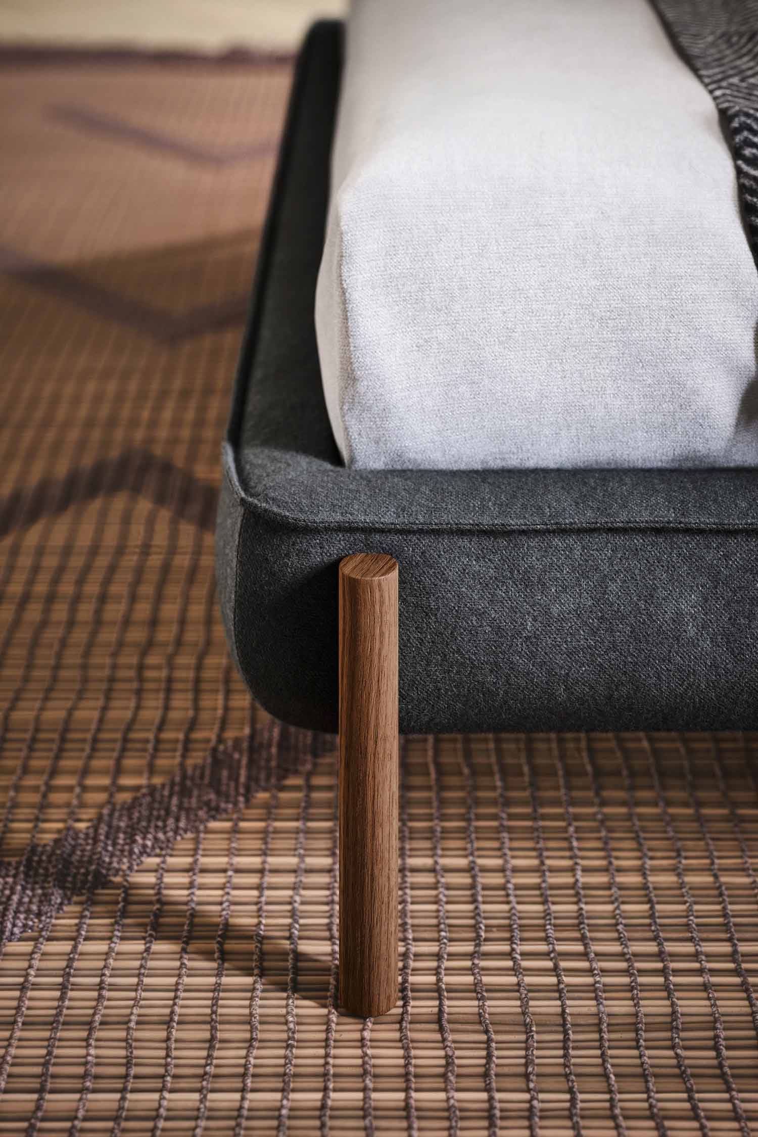 A detailed close-up of the dark gray upholstered bed frame, showing a single, cylindrical wooden leg. The bed rests on a textured, brown woven mat or rug.