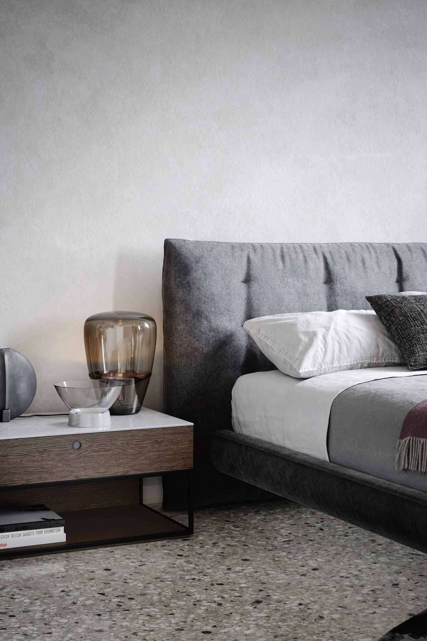 A close-up side view of a gray upholstered bed and tufted headboard with an attached wooden nightstand. The nightstand features a white top and a dark wooden drawer, holding a large, tinted glass table lamp and small accessories.