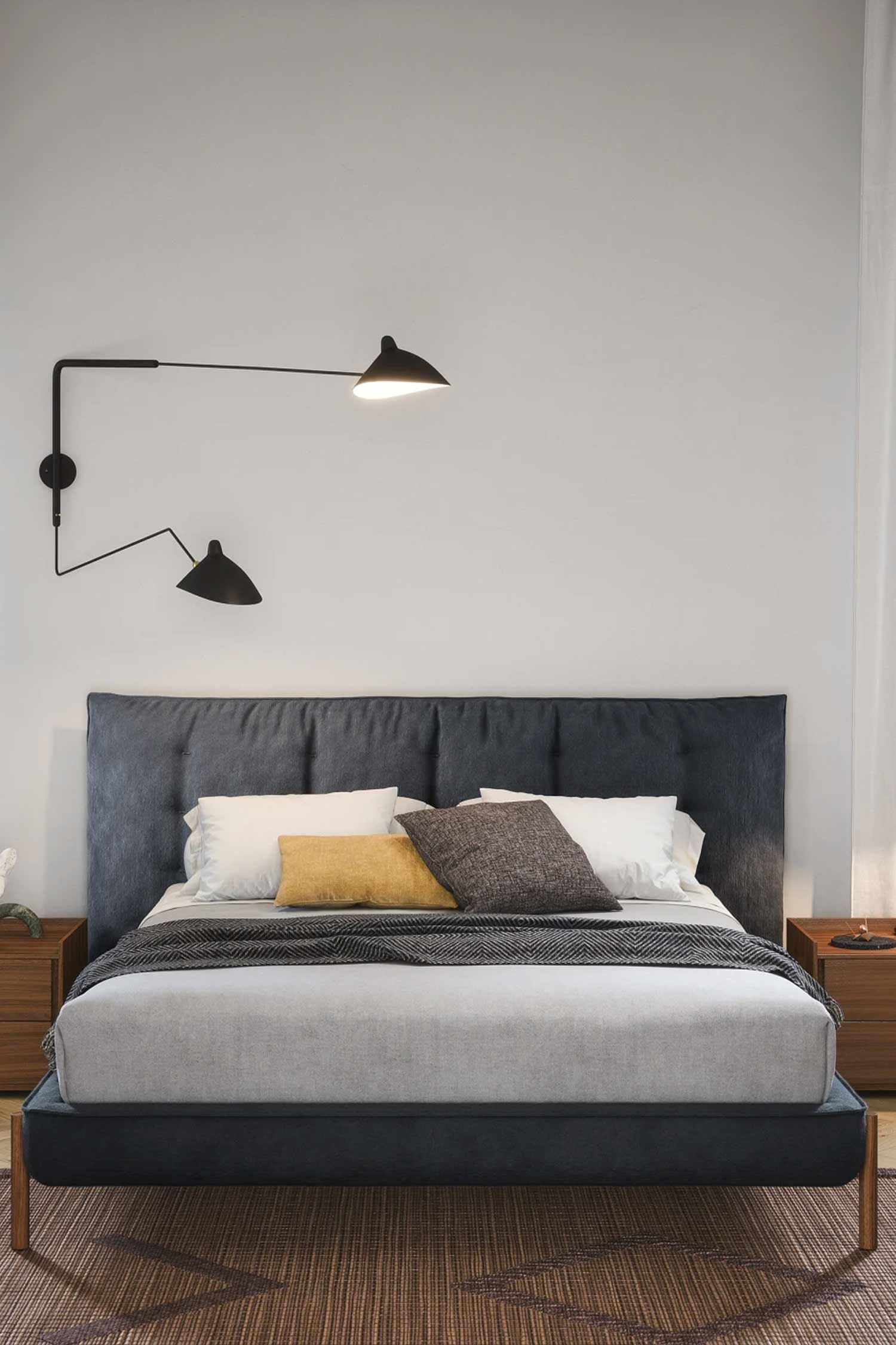 A stylish bedroom featuring a dark blue upholstered bed with a tufted headboard, centered against a white wall. The bed is dressed with a gray blanket and accented with yellow and dark gray pillows. Two adjustable black wall sconces provide light above the bed.