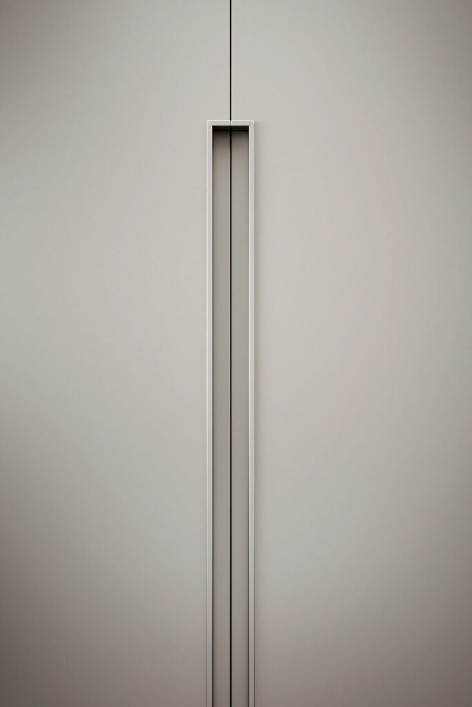 A vertical, close-up view of a minimalist, light gray wardrobe door. The focus is on the long, recessed vertical handle, which is a rectangular, metallic frame that runs from the top to the bottom of the door. The handle has a thin, dark line running down its center, emphasizing the sleek, contemporary design and clean lines of the furniture.