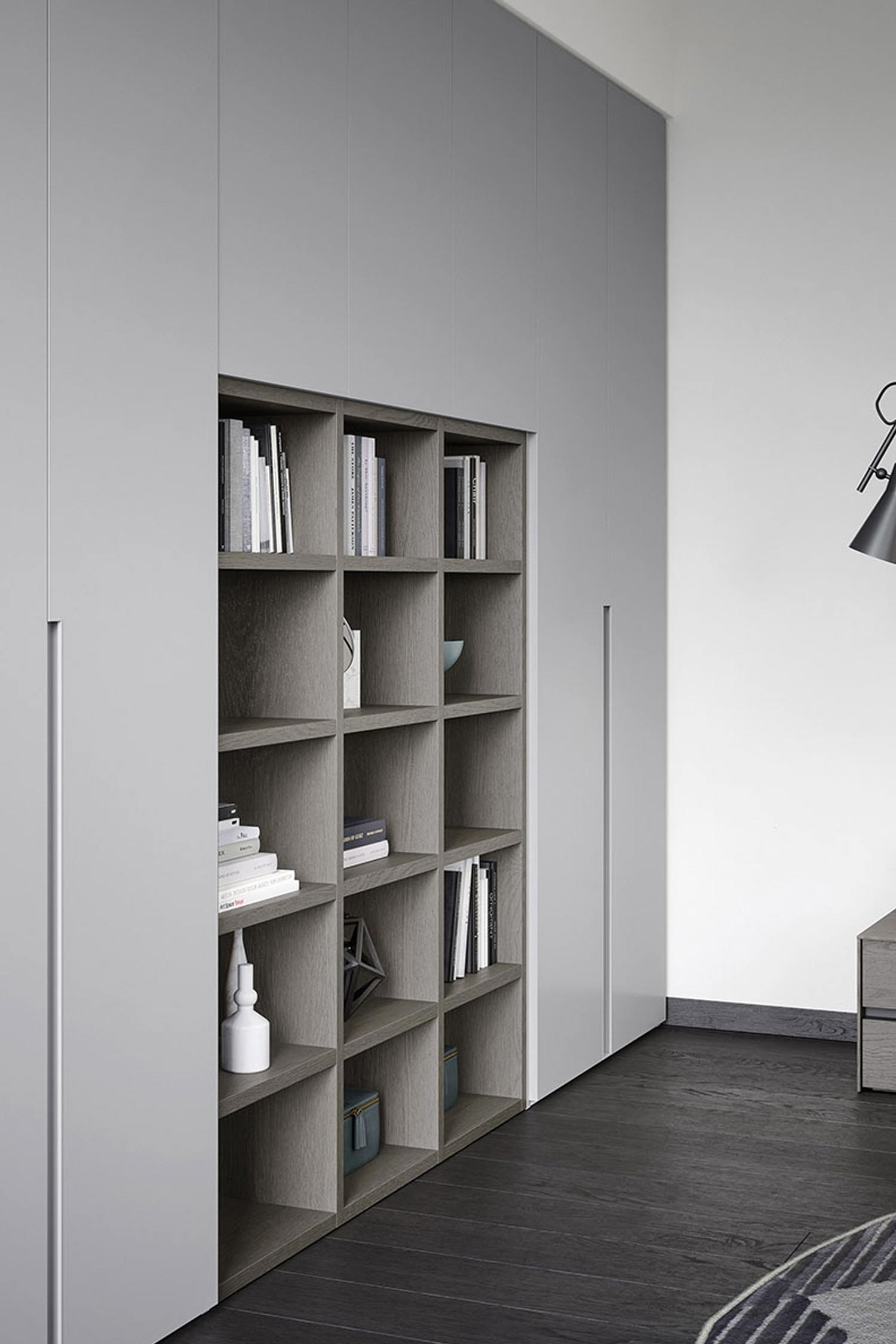 A detailed shot of a minimalist, gray hinged wardrobe system. A large, open shelving unit is built into the center, featuring a grid of square cubbies. The shelves are a darker, textured wood and are filled with a mix of neatly stacked books and various decorative items. The closed wardrobe doors on either side of the bookshelf are a lighter gray with integrated vertical handles. The dark wood floor contrasts with the light gray of the wardrobes, creating a clean, sophisticated look.