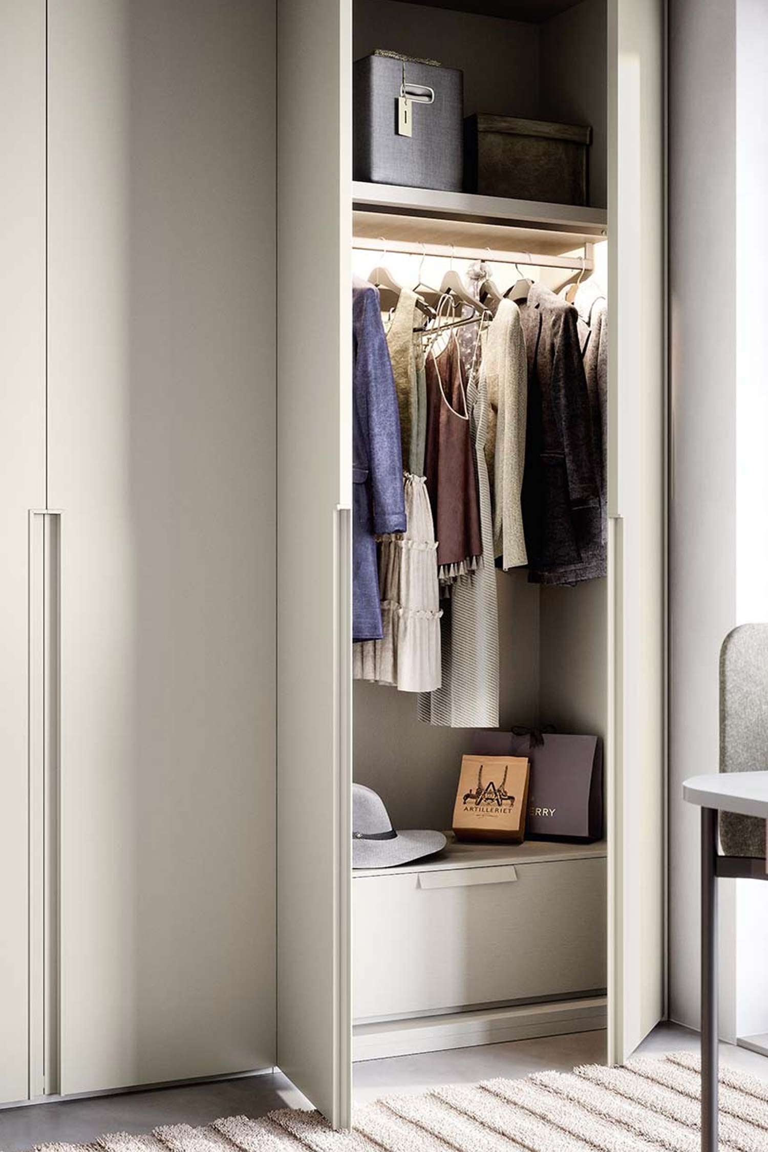 A close-up shot of a modern hinged wardrobe with a light gray, matte finish. One door is partially ajar, revealing a brightly lit interior. The inside features a hanging rod with a mix of clothes and a lower shelf with a gray hat and two shopping bags. Above the clothes, two gray storage boxes are neatly placed on a shelf. The image highlights the clean interior organization and the soft lighting of the wardrobe.