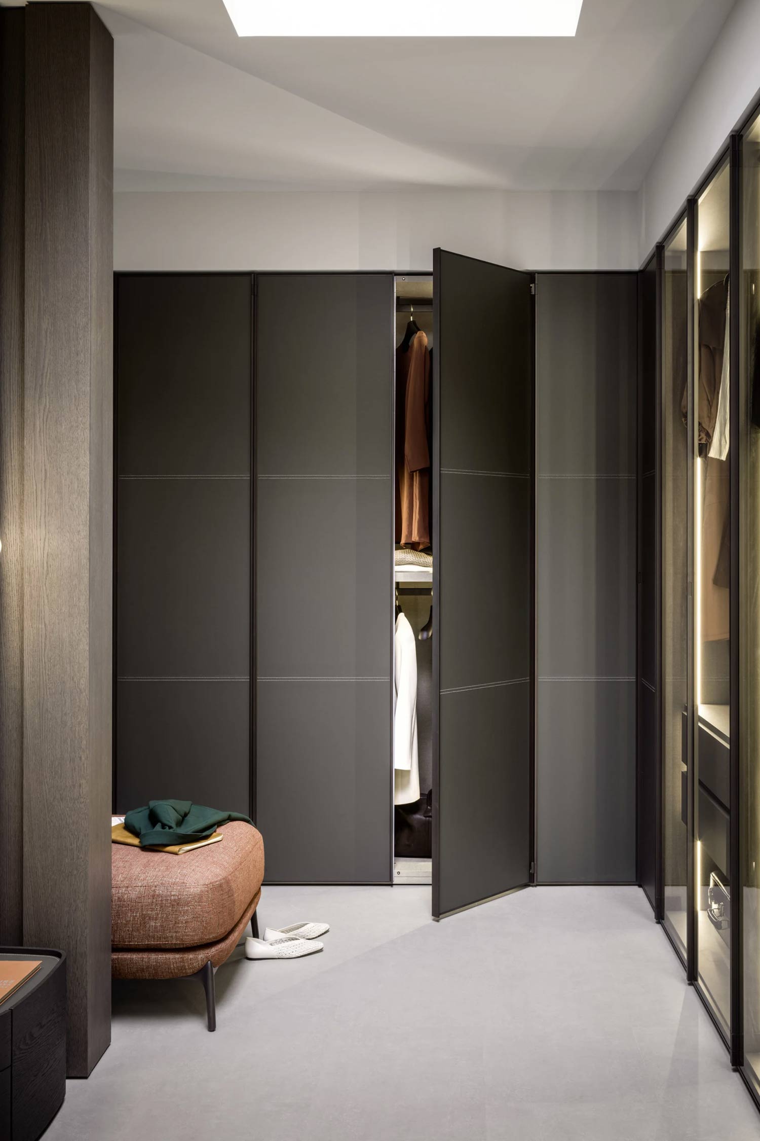 A luxurious walk-in closet or dressing area with a muted color palette. The majority of the back wall is covered by sleek, dark gray floor-to-ceiling wardrobes with subtle horizontal paneling. One set of wardrobe doors on the right is ajar, revealing hanging clothes in warm tones like a brown jacket and a white shirt. To the right, there's a modern glass-fronted closet unit with internal lighting, showcasing more clothing and accessories. In the foreground on the left, a rectangular rust-colored upholstered ottoman with dark legs has a dark green folded garment and a pair of light-colored slippers resting on it. A tall, dark wooden pillar stands to the left of the ottoman, with a hint of a light fixture on its side. The floor is a light gray, possibly concrete or large tiles,
