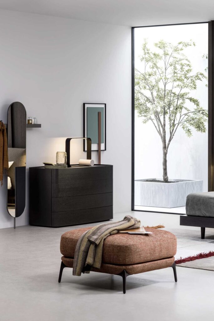 A modern bedroom with minimalist decor. In the foreground, a rust-colored upholstered ottoman with dark metal legs has a striped throw blanket and a book resting on it. To the left, against a white wall, stands a tall, dark wooden mirror with an organic shape. Adjacent to it, a dark wooden dresser with four drawers holds a contemporary black and gold lamp, a glass with a candle, and a few small decorative items. Above the dresser, a small dark shelf is mounted on the wall. A framed abstract art piece leans against the wall behind the dresser. A large, floor-to-ceiling window on the right side of the room offers a view of a minimalist outdoor area with a slender tree planted in a white planter. A corner of a dark bed frame is visible on the far right. The floor is a light, neutral color, possibly concrete or large tiles.