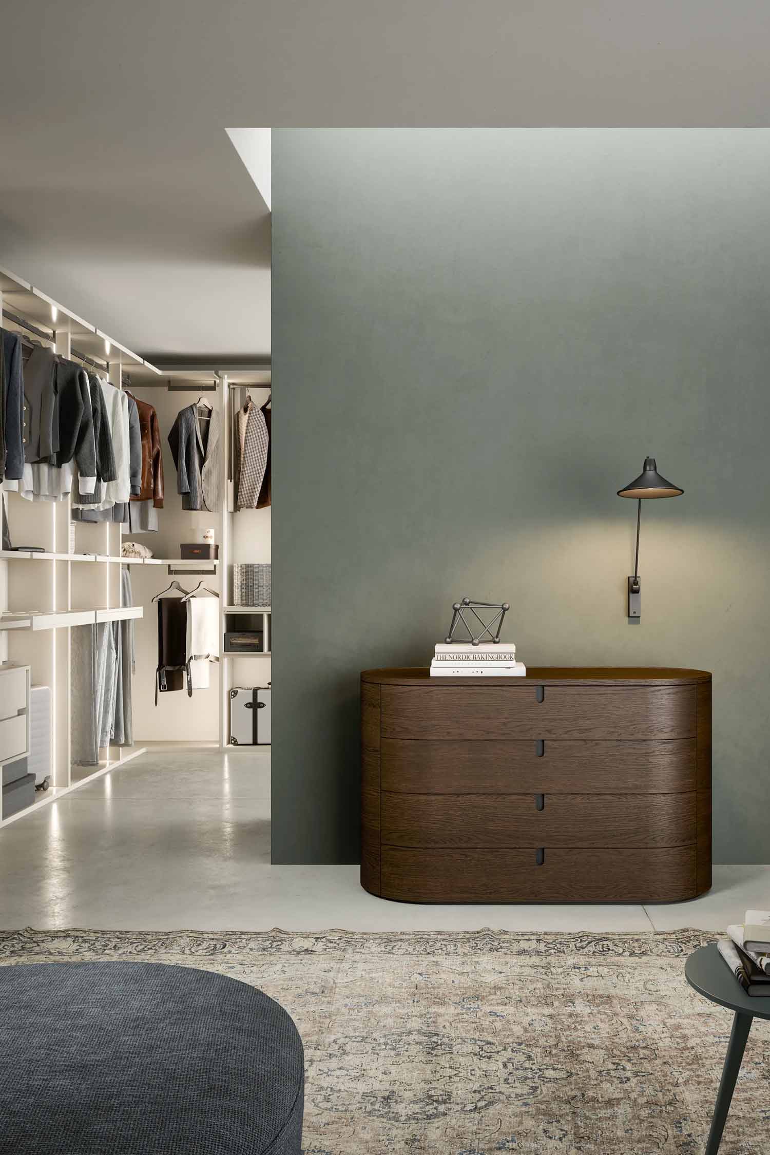 Contemporary dressing area or closet entrance featuring a large, oval, dark wood Vesta luxury four-drawer chest of drawers against a dark sage green wall. A brightly lit walk-in closet is visible in the background.