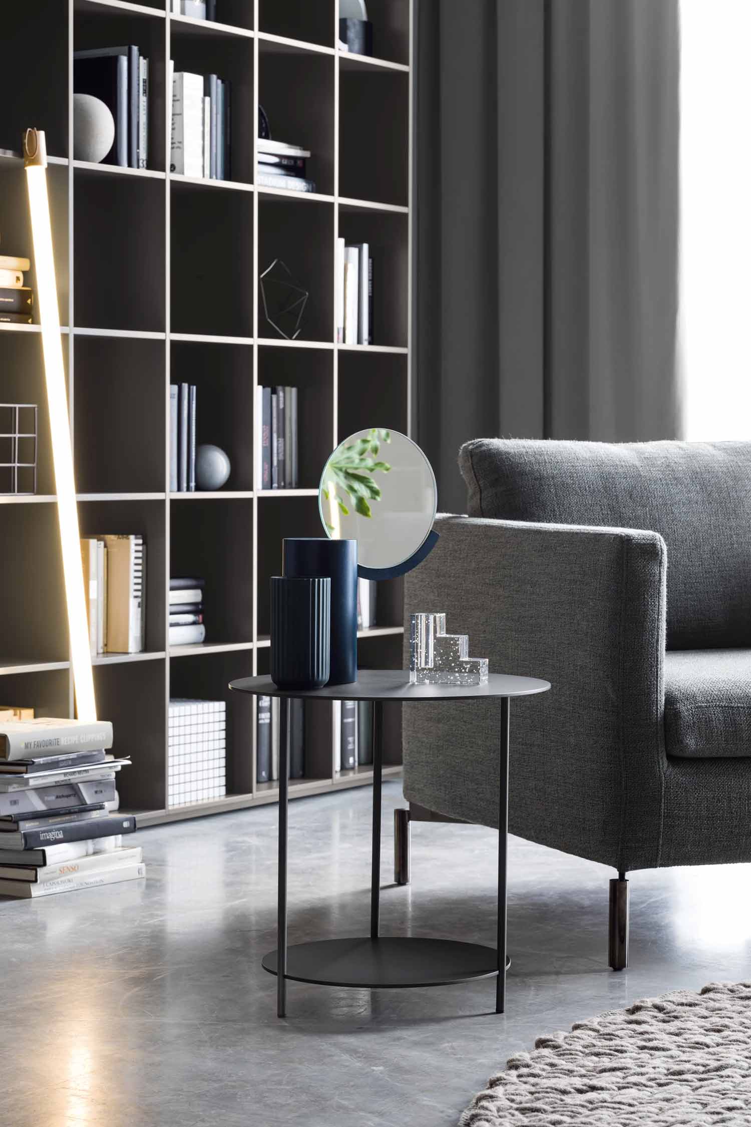 A close-up shot of a modern living room setting. A round, two-tiered black metal side table is positioned next to a gray sofa. On the table, there is a tall, ribbed dark blue vase, a small round mirror with a plant reflected in it, and a stack of clear glass blocks. In the background, a dark gray modular bookcase is filled with books and decor, and a tall, glowing light fixture stands to the left. The floor is a polished light gray, and a gray curtain hangs in the background.
