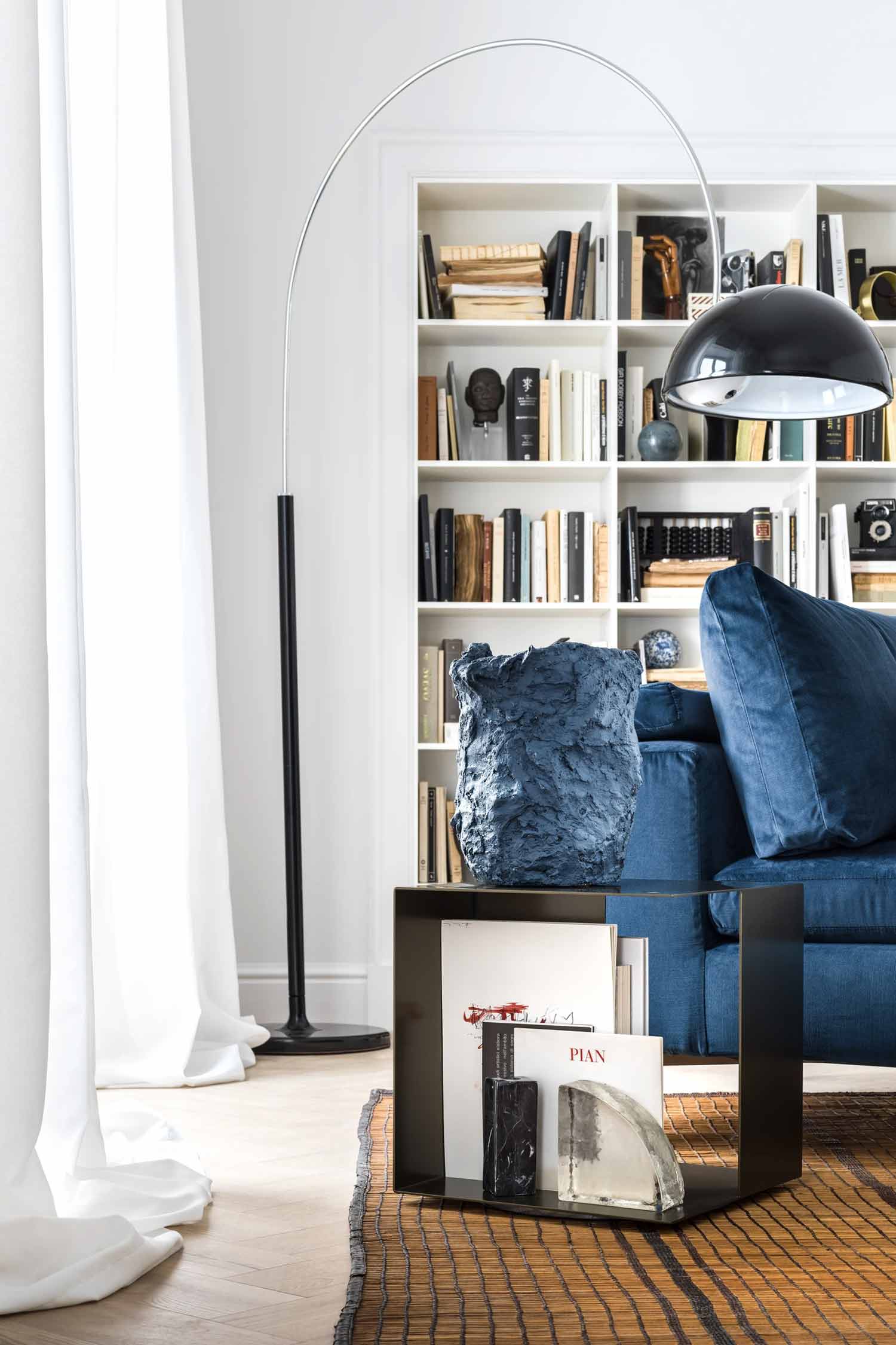 A detail shot focusing on an arched floor lamp and a side table in a living room. The large, black metal lamp curves over a blue velvet sofa. A black, square-framed side table with two tiers holds a textured, sculptural blue vase on top. Books and magazines are neatly stacked on the lower shelf of the table. A white, built-in bookcase is visible in the background, filled with a mix of books and objects. The floor is a light-colored wood, and a textured, striped rug is visible beneath the furniture. Sheer white curtains hang in the background.