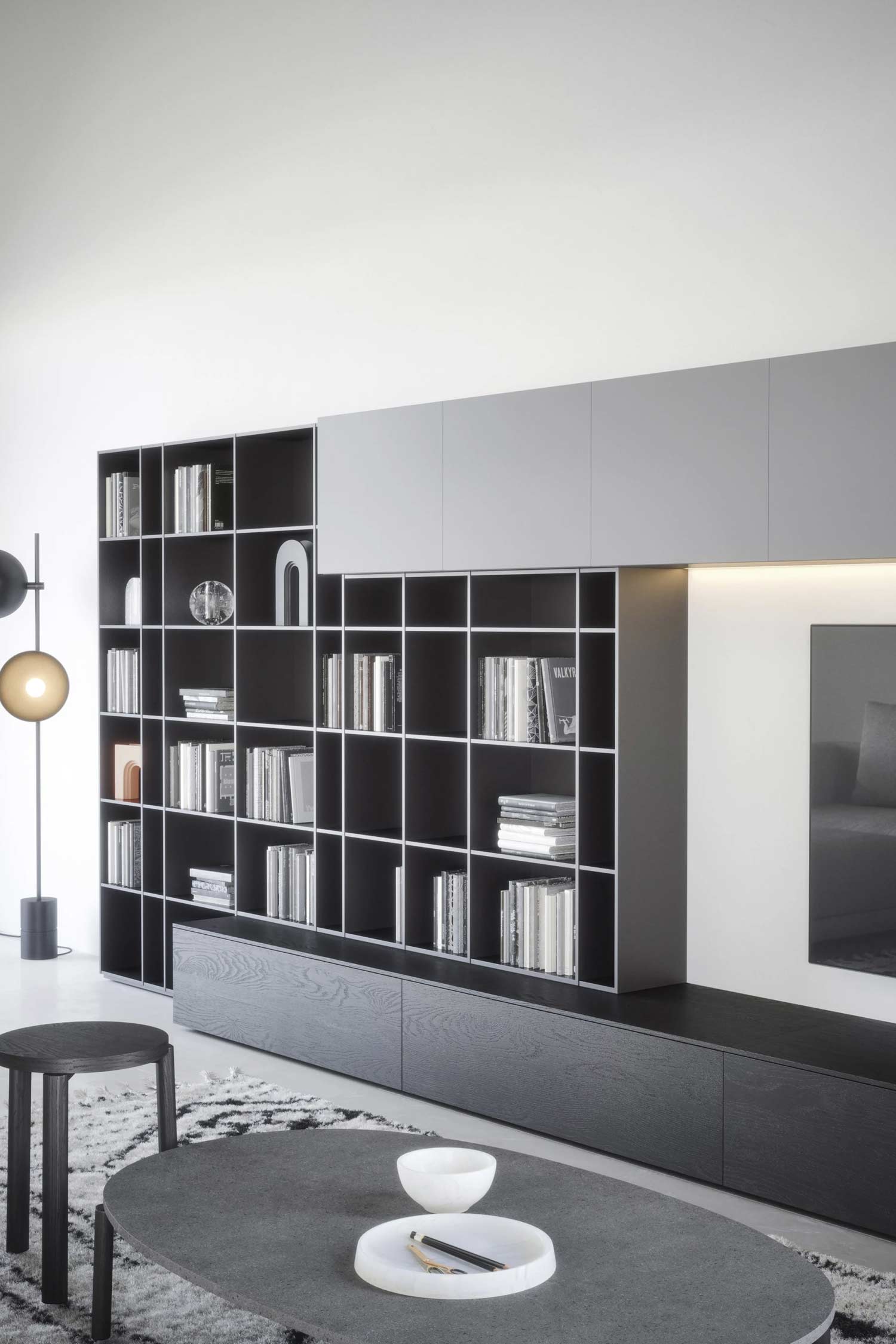 A close-up of a modern, modular wall unit in a living room. The unit consists of a low, horizontal dark wooden cabinet with a grid-like bookcase sitting on top of its left side. The bookcase is dark with thin white dividers, and is filled with books and decor. Above the bookcase and to the right is a floating, light gray storage cabinet. A black television is mounted on the wall to the right. In the foreground, there is a round dark gray coffee table with a small white bowl on it, a round stool to the left, and a shaggy black and white patterned rug.