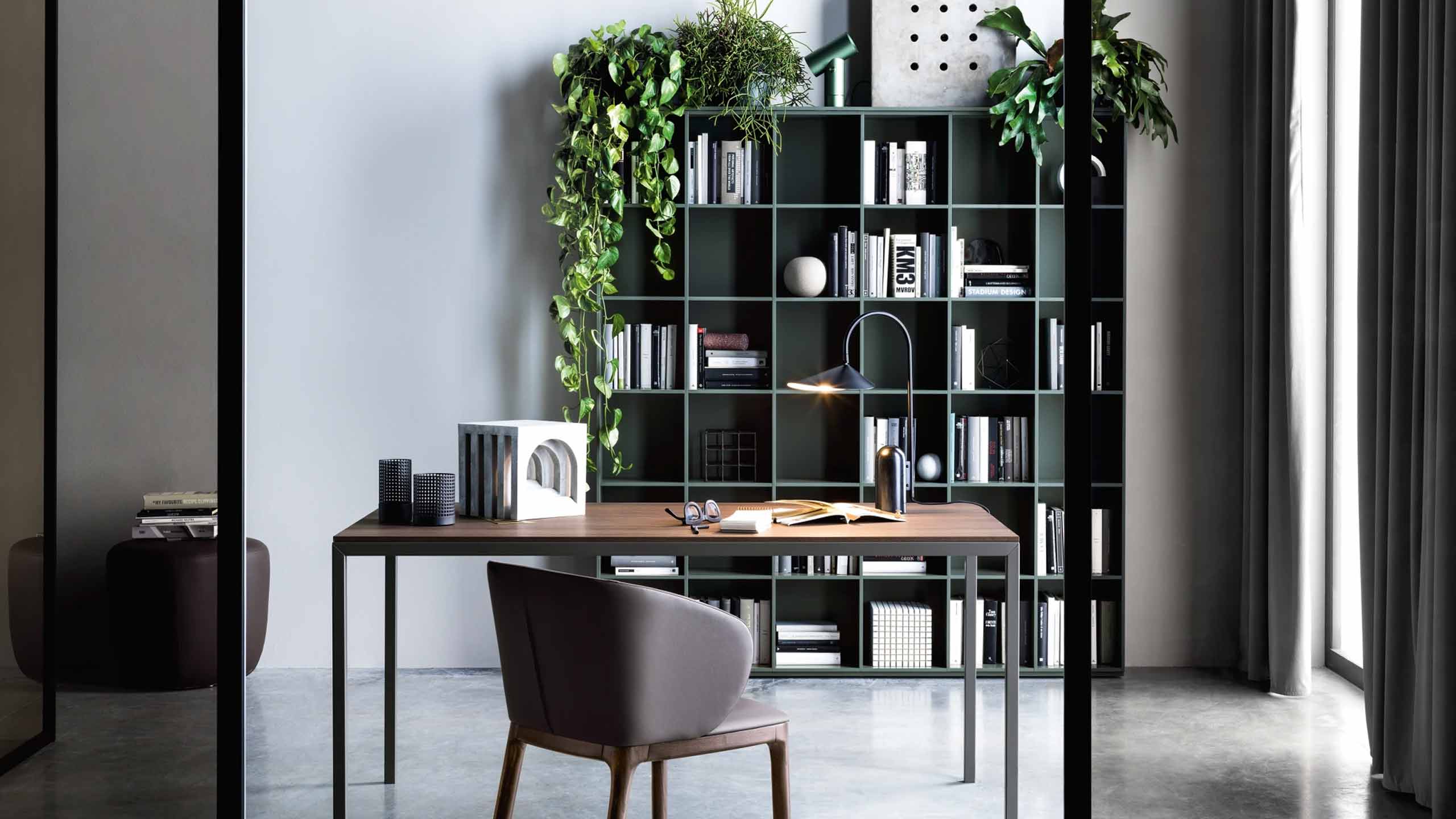 A home office or study with a modern, dark teal modular bookcase. The bookcase is filled with a mix of books, decorative objects, and is adorned with lush green plants trailing from the top. In front of the bookcase is a sleek wooden desk with a metal frame, a gray chair, a task lamp, and various accessories like glasses, a book, and a decorative object. The room has a gray wall, a large window with a sheer curtain, and a glass partition.