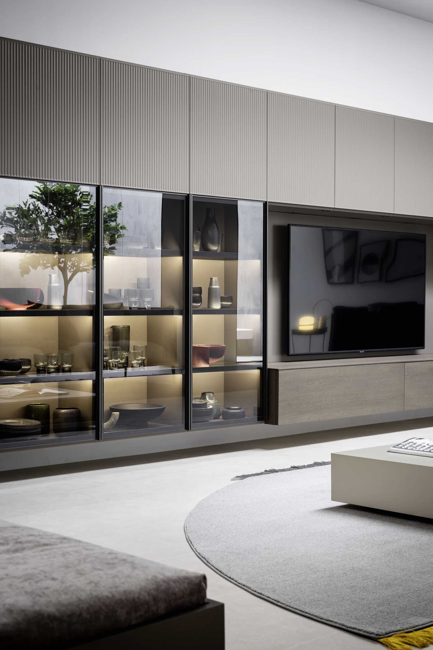 A detailed view of a sophisticated wall unit in a modern living room. The unit is a mix of textures and materials, featuring light brown ribbed cabinet fronts at the top. On the left, four glass-fronted display cases with black frames and internal lighting showcase a collection of light-colored bowls, glassware, and decorative objects on glass shelves. To the right, a television is mounted on a gray panel above a light wood media console. A soft gray rug with a round design is on the light-colored tiled floor, with a part of a sofa visible in the foreground.