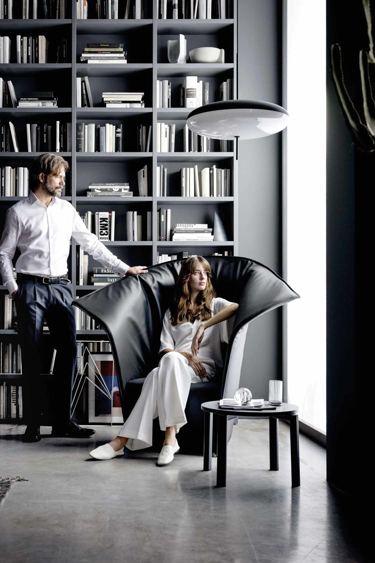 A modern and sophisticated interior featuring a man standing and a woman seated in a unique armchair, surrounded by contemporary design elements. The man, with a beard and dressed in a white shirt and dark trousers, stands to the left, resting his hand on the back of a black, high-backed armchair. The woman, with long hair and wearing a white outfit, is seated comfortably in the armchair, looking towards a tall window on the right. Behind them, a floor-to-ceiling gray bookshelf is densely packed with books. A black round side table with a few items sits next to the armchair. A large, flat pendant light fixture hangs from the ceiling.
