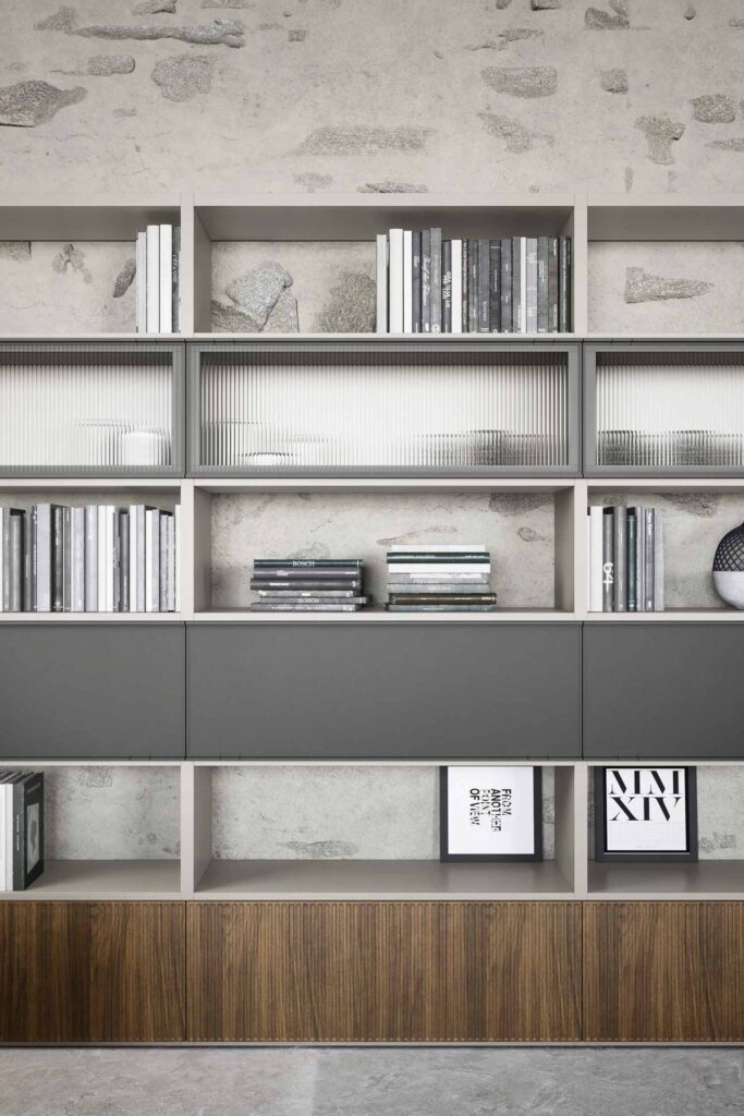 A close-up of a modern, modular shelving unit with a mix of open and closed storage, set against a stone wall background. The unit features open shelves with concrete-like back panels, filled with neatly arranged books, some decorative stones, and a white vase. Three sections have a light-colored, ribbed glass panel. Below these, there are three dark gray, solid-fronted cabinets. The very bottom section of the unit consists of two dark wood panels. Two framed art prints are displayed on the lowest open shelf. The overall design is a blend of rustic and contemporary elements.