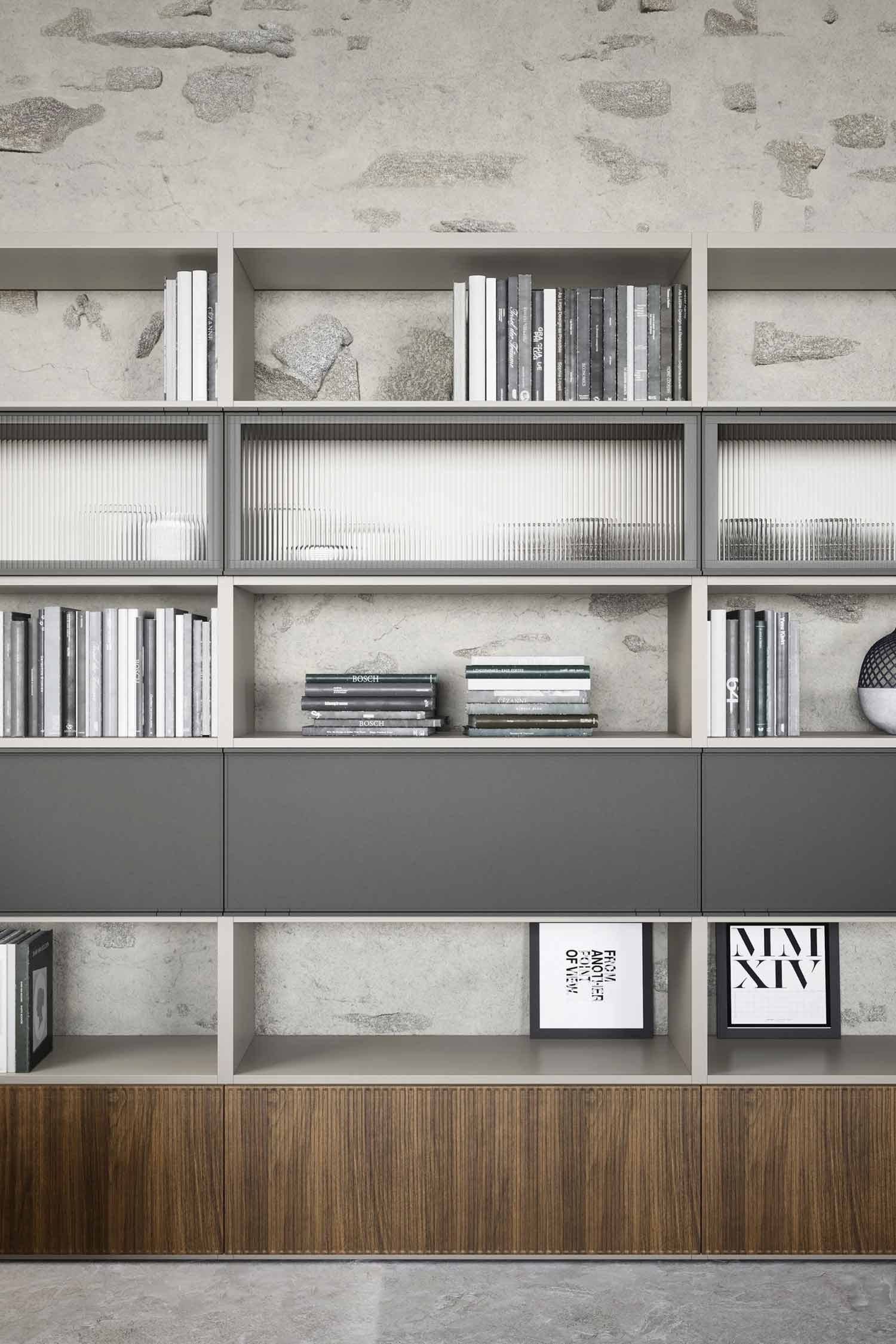 A close-up of a modern, modular shelving unit with a mix of open and closed storage, set against a stone wall background. The unit features open shelves with concrete-like back panels, filled with neatly arranged books, some decorative stones, and a white vase. Three sections have a light-colored, ribbed glass panel. Below these, there are three dark gray, solid-fronted cabinets. The very bottom section of the unit consists of two dark wood panels. Two framed art prints are displayed on the lowest open shelf. The overall design is a blend of rustic and contemporary elements.