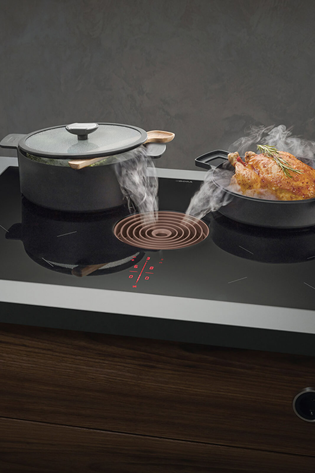 Compact BORA Pure cooktop in matte black with built-in round extractor; two graphite-grey saucepans simmer on adjacent zones, their lids beaded with condensation, emphasising efficient, quiet ventilation in a cosy evening kitchen scene.